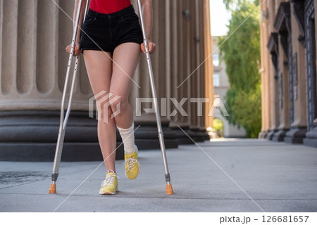 Young red-haired woman in shorts moves with crutches outdoors. Young red-haired woman in shorts moves with crutches outdoors. 126681657