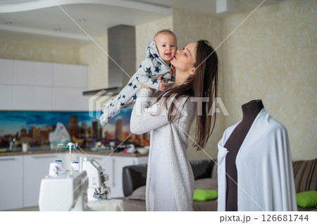 A young woman sews in the kitchen and holds a small child. Mom teaches her little son to sew on a sewing machine. Self-isolation homework 126681744