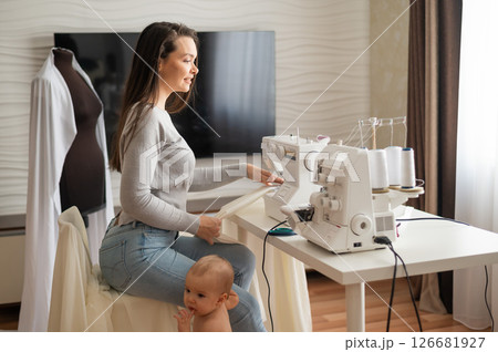Young woman sews at home and holds a small child. Mom teaches her little son to sew on a sewing machine Young woman sews at home and holds a small child. Mom teaches her little son to sew on a sewing machine 126681927
