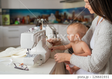 A young woman sews in the kitchen and holds a small child. Mom teaches her little son to sew on a sewing machine. Self-isolation homework 126681931
