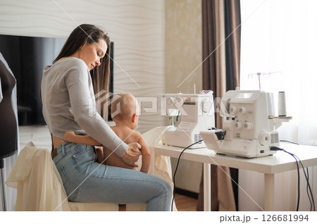 Young woman sews at home and holds a small child. Mom teaches her little son to sew on a sewing machine 126681994