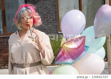 Woman in colored hair walks with an armful of balloons and drinks a refreshing beverage Woman in colored hair walks with an armful of balloons and drinks a refreshing beverage 126682107