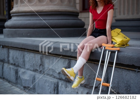 Young red-haired woman in shorts moves with crutches outdoors. 126682182