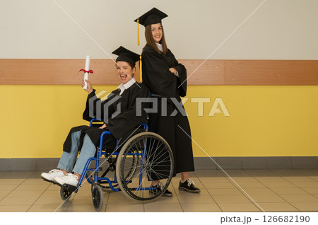Happy young woman and woman in wheelchair in graduate gown with diploma in hands. Inclusive education. 126682190