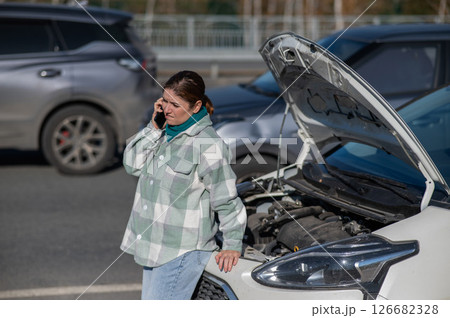 Caucasian woman calls on the phone and asks for help while standing near a car with an open hood.  126682328