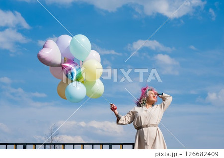 A woman with colored hair with an armful of balloons against a blue sky. 126682409