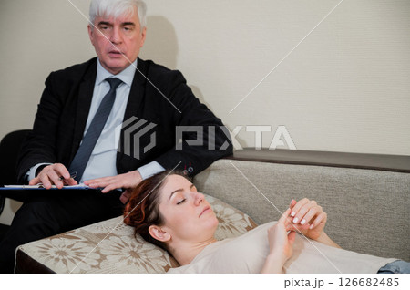 Caucasian woman lies on a couch at a reception with a psychotherapist. An elderly man works as a psychiatrist Caucasian woman lies on a couch at a reception with a psychotherapist. An elderly man works as a psychiatrist 126682485