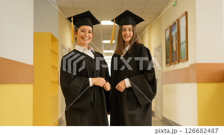 Two young women in graduation gowns stand in a university corridor.  126682487