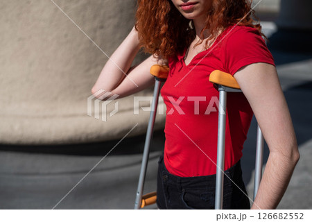 Young red-haired woman in shorts moves with crutches outdoors. Young red-haired woman in shorts moves with crutches outdoors. 126682552