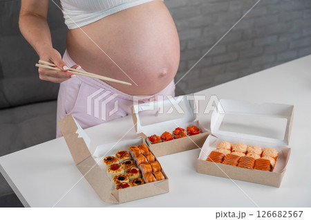 A pregnant woman holds chopsticks and stands at the table with rolls. Close-up of the belly. 126682567