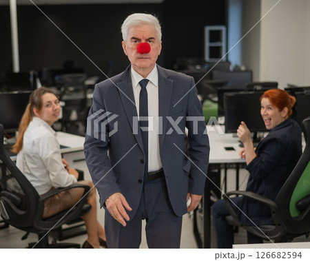 Two Caucasian women look at a serious elderly man with a clown nose in the office. Two Caucasian women look at a serious elderly man with a clown nose in the office. 126682594