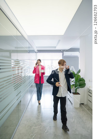 Two colleagues are walking through the office and talking on the phones.  126682703