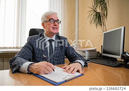 A confident elderly gray-haired man in a checkered suit and glasses sits at his desk in the office. A confident elderly gray-haired man in a checkered suit and glasses sits at his desk in the office. 126682724