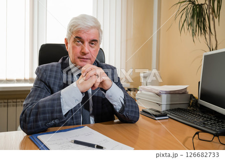 Confident elderly gray-haired man in a plaid suit sits at his desk in the office Confident elderly gray-haired man in a plaid suit sits at his desk in the office 126682733