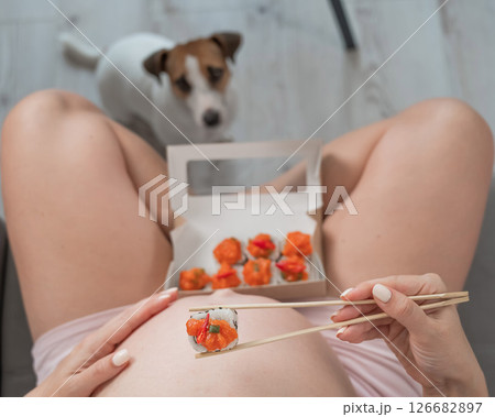 A pregnant woman sits on the sofa and eats rolls. Jack Russell Terrier dog sits on the floor and begs for food from its owner.  126682897