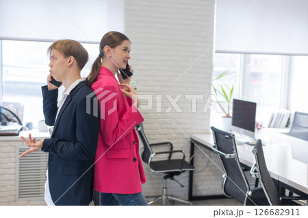 Two colleagues stand back to back and talk on their phones. Two colleagues stand back to back and talk on their phones. 126682911
