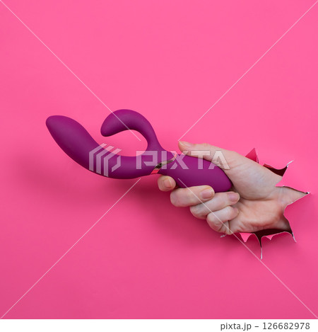 Female hand sticking out with raspberry sex toy from pink paper background. Female hand sticking out with raspberry sex toy from pink paper background. 126682978