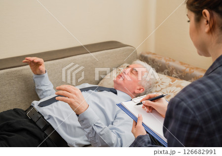 An elderly man lies on a couch and holds his hands hanging during an appointment with a hypnotherapist. An elderly male patient in an individual consultation with a female psychologist An elderly man lies on a couch and holds his hands hanging during an appointment with a hypnotherapist. An elderly male patient in an individual consultation with a female psychologist 126682991
