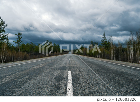 Expansive highway stretches into distance beneath dramatic stormy sky with scattered trees along the roadside Expansive highway stretches into distance beneath dramatic stormy sky with scattered trees along the roadside 126683820