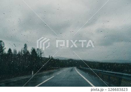 Raindrops cling to the windshield, blurring the view of an empty road winding through a gray, overcast landscape Raindrops cling to the windshield, blurring the view of an empty road winding through a gray, overcast landscape 126683823