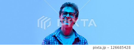 Happy young guy in glasses wearing checkered shirt laughing with closed eyes, looking cheerful and carefree in neon light against gradient background Happy young guy in glasses wearing checkered shirt laughing with closed eyes, looking cheerful and carefree in neon light against gradient background 126685428