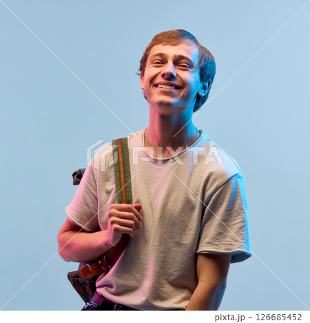 Smiling boy with backpack standing in neon light against gradient background showing happy mood and relaxed feeling. Open heart and light joy 126685452