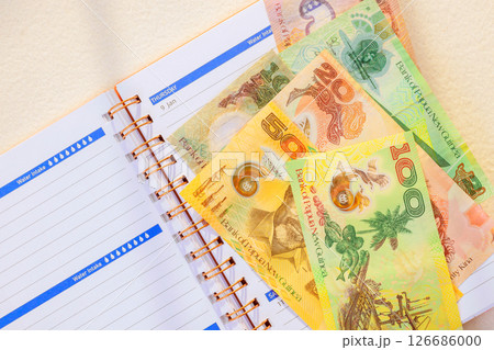 Various denominations Papua New Guinea banknotes lay atop an open planner with date noted for scheduling purposes. Various denominations Papua New Guinea banknotes lay atop an open planner with date noted for scheduling purposes. 126686000