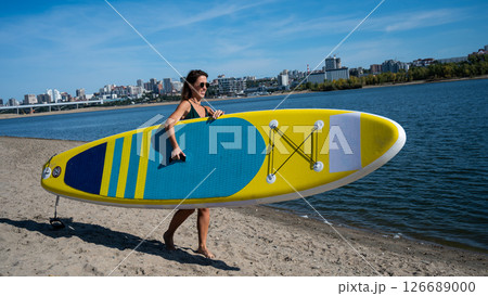 Caucasian woman walks along the beach and carries a sup board on the river in the city. Summer sport. Caucasian woman walks along the beach and carries a sup board on the river in the city. Summer sport. 126689000