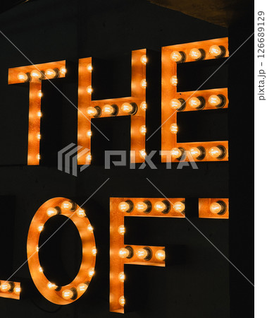 Glowing capital letters spelling The Loft emitting warm light, casting soft illumination against shadowy background, creating inviting nighttime signage atmosphere. Large illuminated letters The Loft Glowing capital letters spelling The Loft emitting warm light, casting soft illumination against shadowy background, creating inviting nighttime signage atmosphere. Large illuminated letters The Loft 126689129