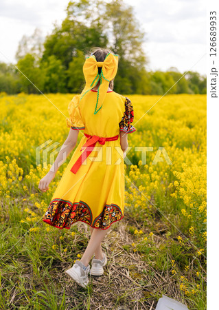 Back view of unrecognizable child girl in yellow dress with traditional Russian pattern, standing posing in field of yellow flowers during sunny summer day, enjoying beauty of nature and warm weather. 126689933