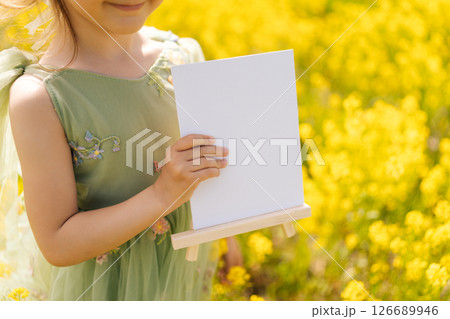 Cropped shot of smiling child girl in green dress holding blank canvas on wooden easel, surrounded by vibrant yellow rapeseed flowers in sunny field, offering copy space for creative designs. 126689946