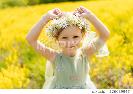 Pretty preschooler girl wearing beautiful flower crown and fairy dress playing in blooming yellow meadow on sunny summer day, enjoying warm weather and vibrant colors of nature, looking at camera. 126689947