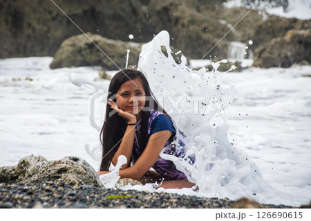 A woman smiles as she sits on a rocky shore when a foaming ocean wave suddenly hits her head A woman smiles as she sits on a rocky shore when a foaming ocean wave suddenly hits her head 126690615