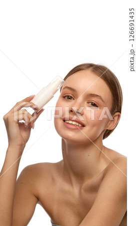 Young woman smiling while holding white cosmetic bottle near forehead, show clean healthy skin and natural makeup against white studio background. 126691435