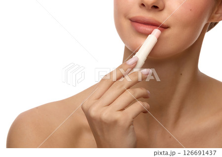 Close-up of woman applying moisturizing balm on lips with healthy skin, gentle smile and light glow on face against white background 126691437