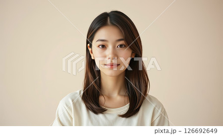 Asian woman with long black hair wearing a plain white t-shirt poses confidently against a soft beige background, showcasing natural beauty and simplicity in portrait photography 126692649