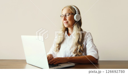 Happy mature woman listening to music in headphones working with laptop sitting at desk at home Happy mature woman listening to music in headphones working with laptop sitting at desk at home 126692780