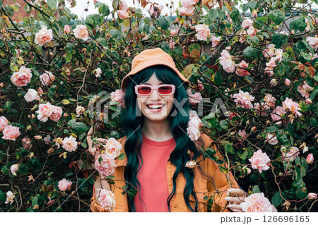 Young laughing hipster woman in bright outfit, bucket hat and sunglasses posing on blooming roses bush background. Urban street fashion. Gen Z. Summer vacation, fun, positive mood vibes 126696165