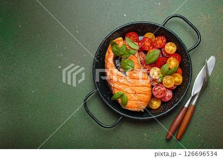 Grilled salmon steak with cherry tomatoes and basil leaves in black pan with cutlery top view on dark green background, space for text 126696534