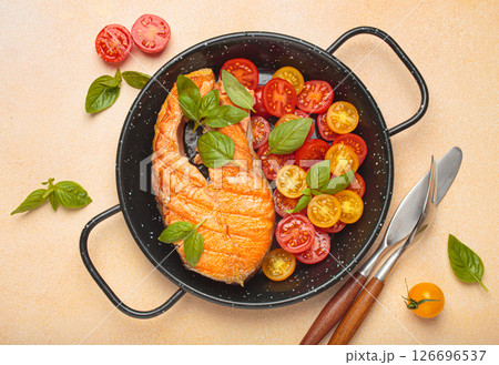 Grilled salmon steak with cherry tomatoes and basil leaves in black pan with cutlery top view on light beige background 126696537