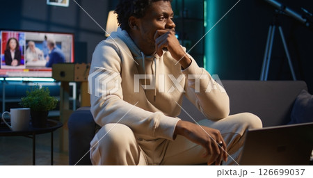 African american male remote worker sitting at home on couch late at night, overthinking about problems. Young man struggling to finish work project before deadline, finding solutions. Camera A. African american male remote worker sitting at home on couch late at night, overthinking about problems. Young man struggling to finish work project before deadline, finding solutions. Camera A. 126699037