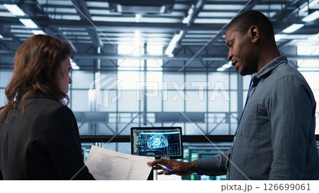 Team of IT professionals looking over data center checkup paperwork, using AI tech on tablet. Colleagues collaborating in server farm, reading documentation, using artificial intelligence, camera B 126699061