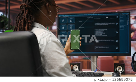 Woman uses green screen phone, doing videocall in front of PC screen with AI LLM chatbot answering prompt. Person uses large language model program during teleconference on mockup smartphone, camera B 126699079