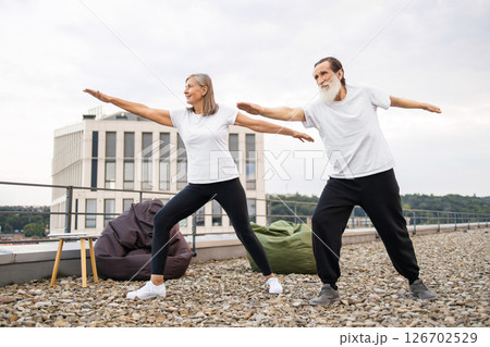Elderly man and woman performing yoga together on rooftop in warrior pose, promoting well-being and fitness Elderly man and woman performing yoga together on rooftop in warrior pose, promoting well-being and fitness 126702529