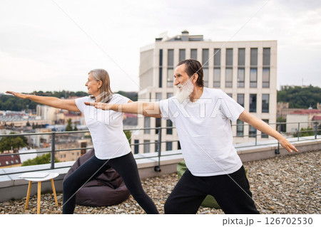 Senior man and woman practicing yoga in outdoor rooftop session overlooking urban landscape. Both wearing casual tops and comfortable clothing while performing warrior yoga poses, emphasizing balance. Senior man and woman practicing yoga in outdoor rooftop session overlooking urban landscape. Both wearing casual tops and comfortable clothing while performing warrior yoga poses, emphasizing balance. 126702530
