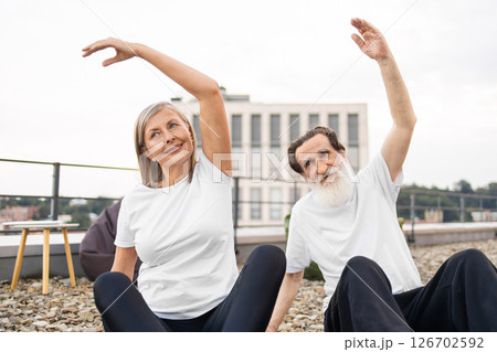 Senior man and woman practicing yoga together outdoors on terrace. Models are elderly white man with beard and elderly Caucasian woman, both wearing white t-shirts and smiling while stretching arms. 126702592