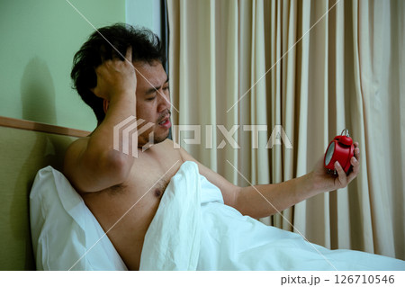 Shirtless man sitting in bed under white blanket looking frustrated while holding red alarm clock in hand Shirtless man sitting in bed under white blanket looking frustrated while holding red alarm clock in hand 126710546