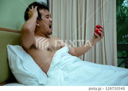 Angry man wakes up in bed, yelling at a red alarm clock in his hand, overwhelmed by the early morning wake-up 126710548