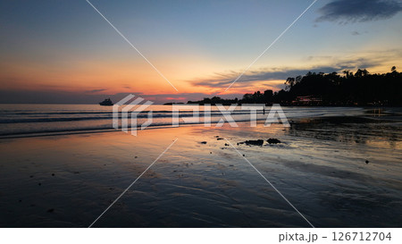 Vibrant sunset reflecting on wet sand with yacht sailing on calm sea Vibrant sunset reflecting on wet sand with yacht sailing on calm sea 126712704