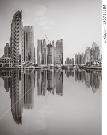 Monochrome city skyline with tall skyscrapers reflecting on calm water under a cloudy sky, Dubai Marina bay UAE 126713194